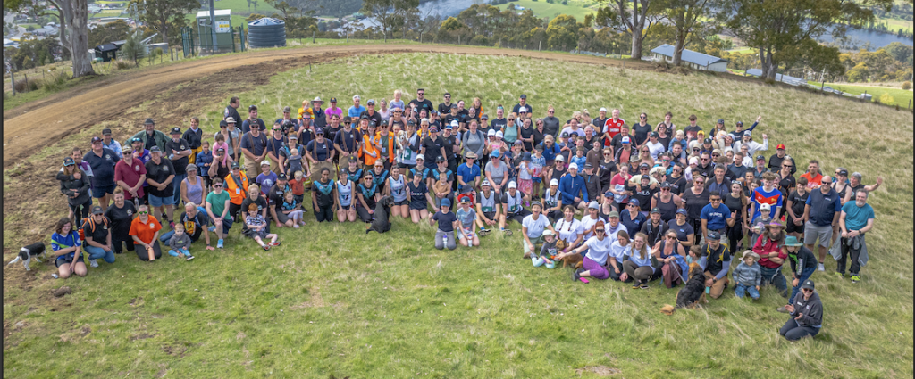 A large group of people pose together outdoors on a grassy hill, surrounded by trees and scenic views of fields and water in the background. The group appears to be participating in an event or gathering.